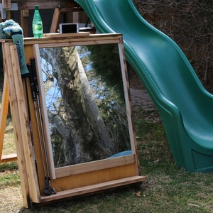 Puede incluir: Un espejo de pr&aacute;ctica de swing de golf de madera con un estante para almacenamiento. El espejo est&aacute; apoyado en un caballete de madera y tiene un guante de golf verde y palos de golf colgados en el lateral. El espejo refleja un &aacute;rbol y un cielo azul.