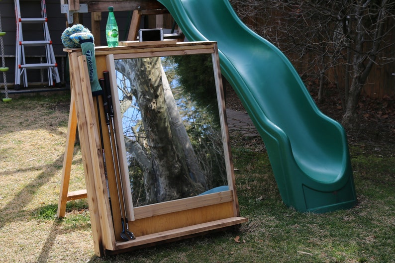 Puede incluir: Un espejo de pr&aacute;ctica de swing de golf de madera con un estante para almacenamiento. El espejo est&aacute; apoyado en un caballete de madera y tiene un guante de golf verde y palos de golf colgados en el lateral. El espejo refleja un &aacute;rbol y un cielo azul.