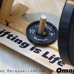 May include: A wooden weightlifting stand with two black weight plates. The smaller plate has the text "SPORTS" and "45 LBS" printed on it. The larger plate has the text "ROGUE" printed on it. The stand has a wooden sign with the text "Lifting is Life" printed on it.