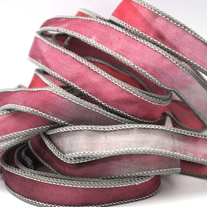 Op de afbeelding: Een stapel roze en grijze lint met een zilveren rand. Het lint is ombre, van een donkerder roze naar een lichter roze.