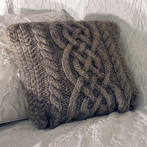 Puede incluir: Una almohada de punto marrón con un patrón de punto de cable. La almohada está sobre una superficie blanca texturizada.