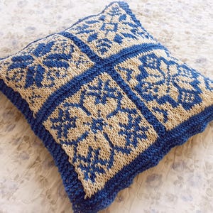 Op de afbeelding: Een vierkant, gebreid kussen met een blauw en beige bloemmotief. Het kussen heeft een herhalend ontwerp van gestileerde bloemen en bladeren in een raster, met een effen blauwe rand en een geschulpte rand.