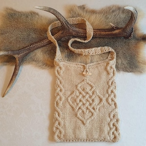 Puede incluir: Un bolso de punto beige con un diseño de nudo celta. El bolso tiene una correa larga y un cierre de botón de madera. El bolso se muestra sobre un fondo de piel sintética.