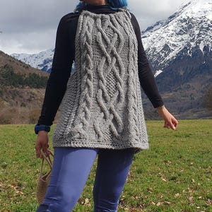 May include: A gray cable-knit tunic with a high neckline. The sleeveless garment features a textured pattern of interwoven cables. The tunic is worn over a black long-sleeve shirt and blue leggings. A small beige bag is held in the hand.