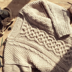 Peut inclure: Un pull beige en tricot torsadé avec un motif texturé et un motif torsadé décoratif. Le pull est présenté sur une surface en bois avec des aiguilles à tricoter, de la laine et des ciseaux.