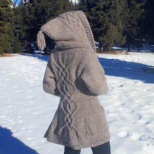 May include: A beige hooded sweater coat with a cable knit design down the back. The coat has a long hood with a tassel and is worn over black trousers in a snowy outdoor setting.