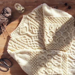 Peut inclure: Couverture en tricot crème avec des motifs de nœuds celtiques et de câbles. Des aiguilles à tricoter, de la laine, des ciseaux et un bougeoir sont disposés autour de la couverture, suggérant un thème artisanal ou de décoration intérieure.