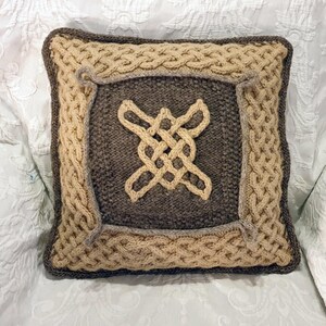 May include: Square knitted pillow with a Celtic knot design. The pillow features a central dark gray square with a light tan knot, framed by a light tan cable knit border and a dark gray edge.