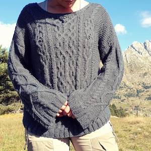 May include: A dark gray, cable-knit sweater with a wide neckline and long sleeves. The sweater features intricate cable patterns on the front and sleeves. The person is wearing khaki pants. The background includes a mountain range and a blue sky.