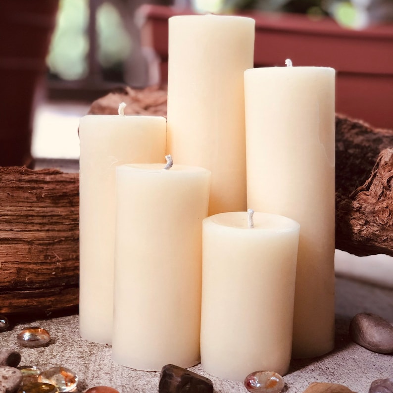 May include: Five ivory-colored pillar candles of varying heights, arranged on a textured brown surface. The candles are all unscented and have a smooth, polished finish.