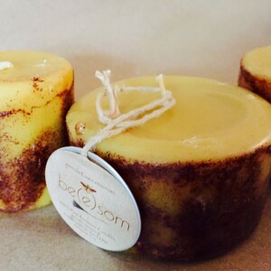 May include: Three yellow candles with brown flecks, two are round and one is square. The square candle has a string tied around it and a label that says "be(e)som" with a bee graphic.