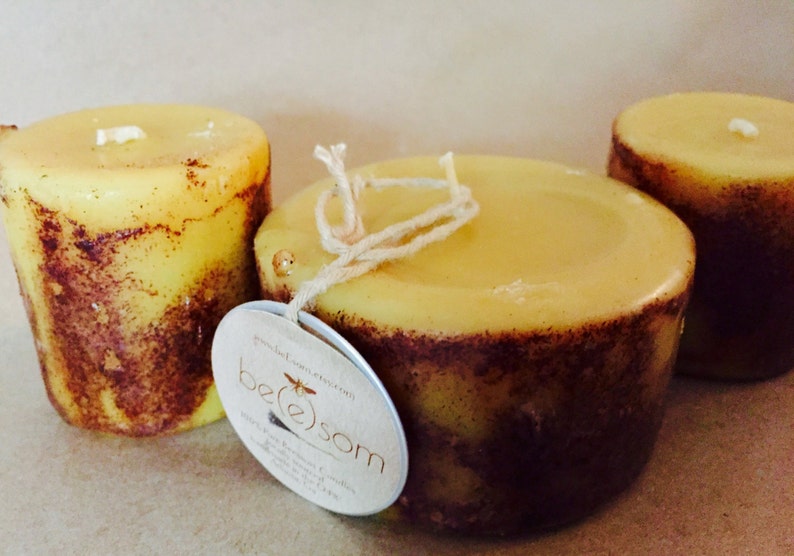 May include: Three yellow candles with brown flecks, two are round and one is square. The square candle has a string tied around it and a label that says "be(e)som" with a bee graphic.