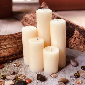 May include: Five ivory pillar candles of varying heights arranged on a piece of driftwood. The candles are on a light gray surface with small decorative stones.
