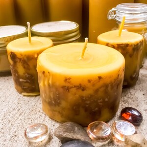 May include: Three yellow beeswax candles with brown marbling. The candles are sitting on a light brown surface with small stones and glass beads.