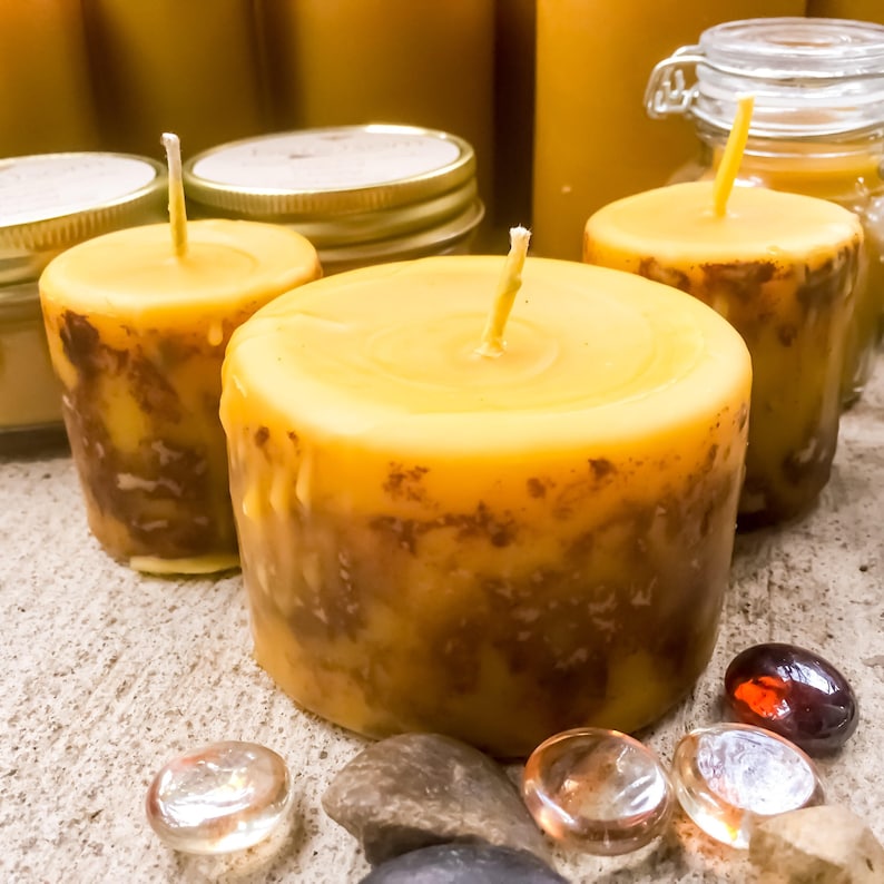 May include: Three yellow beeswax candles with brown marbling. The candles are sitting on a light brown surface with small stones and glass beads.