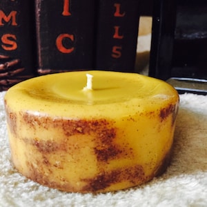 May include: A round, yellow candle with a brown speckled design. The candle has a single wick and is sitting on a white surface.