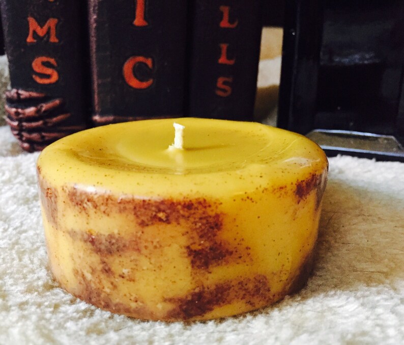 May include: A round, yellow candle with a brown speckled design. The candle has a single wick and is sitting on a white surface.