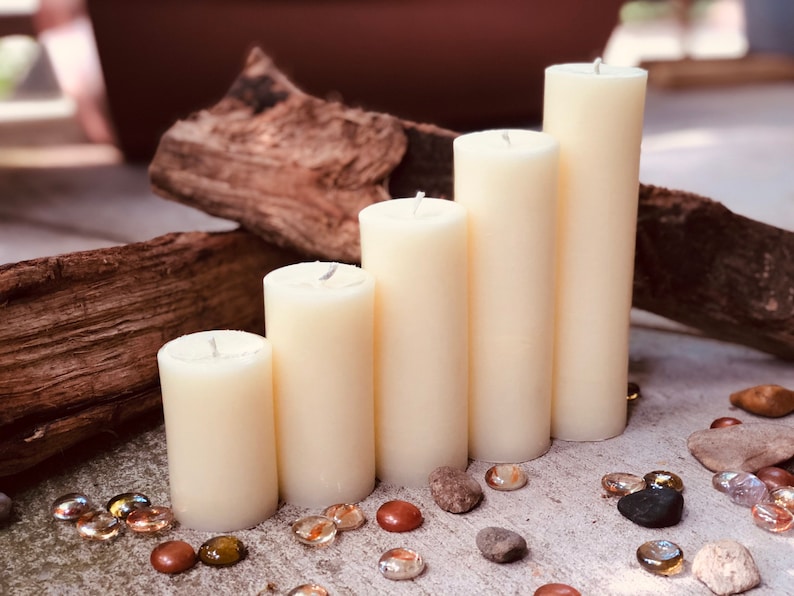May include: Five ivory pillar candles of varying heights arranged on a rustic wooden surface with small decorative stones scattered around them.