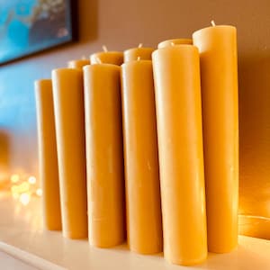 May include: A collection of cylindrical, golden-yellow pillar candles. The candles are arranged closely together, standing upright on a white surface. Each candle has a small, white wick at the top. The background is a warm, neutral color.