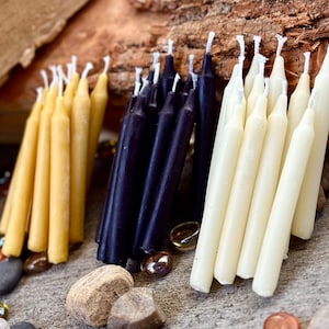 May include: A collection of three types of candles: white, black, and yellow. The candles are arranged in a fan shape and are all the same size. The candles are on a wooden surface.
