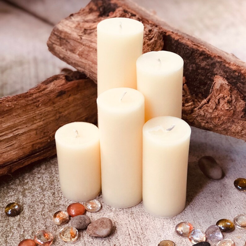 May include: Five ivory pillar candles of varying heights arranged in a pyramid shape on a rustic wooden surface. The candles are surrounded by small decorative stones.