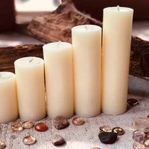 May include: Five ivory pillar candles of varying heights arranged in a row on a light wood surface with small decorative stones scattered around them.