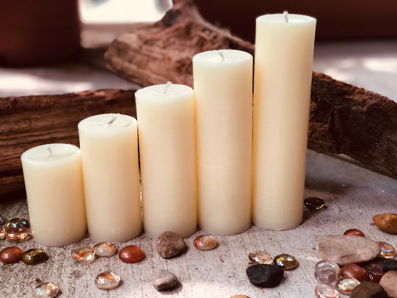 May include: Five ivory pillar candles of varying heights arranged in a row on a light wood surface with small decorative stones scattered around them.