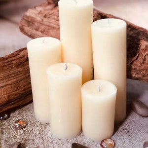 May include: Five ivory pillar candles of varying heights arranged on a rustic wooden surface. The candles are surrounded by small decorative stones.