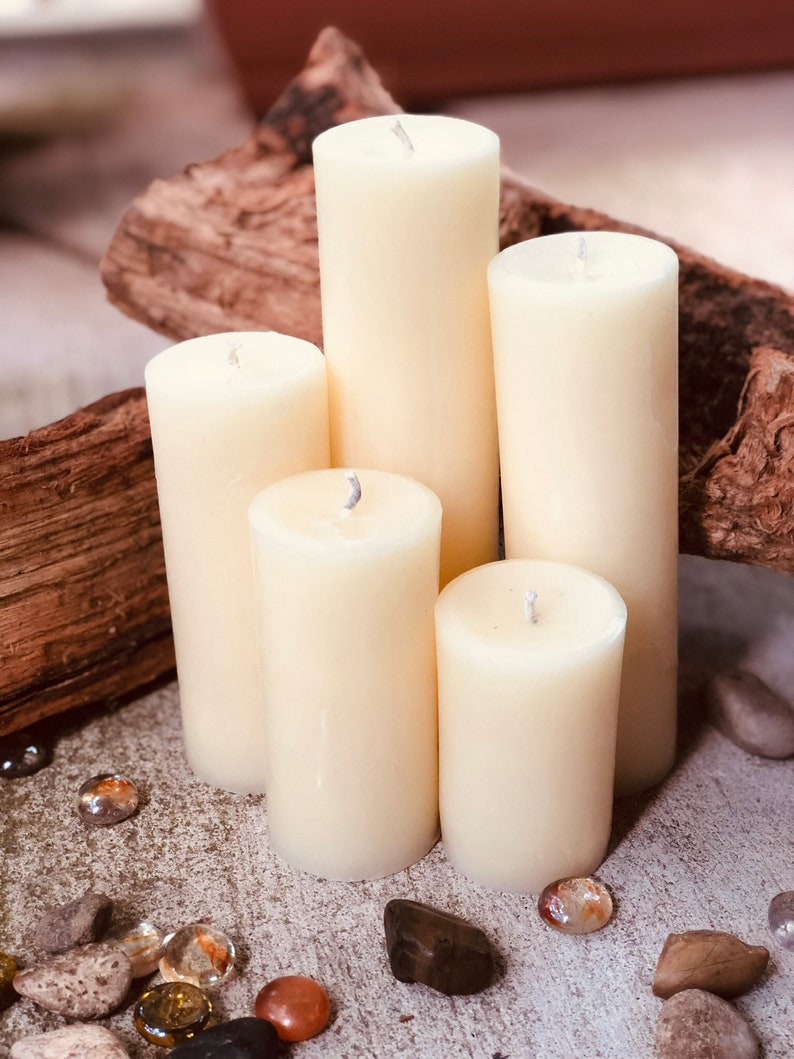 May include: Five ivory pillar candles of varying heights arranged on a rustic wooden surface. The candles are surrounded by small decorative stones.