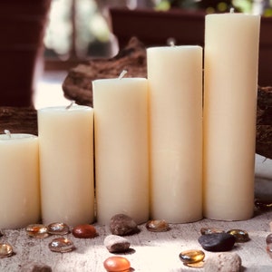 May include: Five ivory pillar candles of varying heights arranged in a staggered formation on a wooden surface. The candles are surrounded by small, clear glass stones.