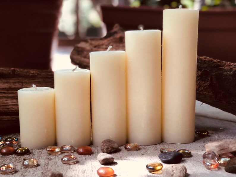 May include: Five ivory pillar candles of varying heights arranged in a staggered formation on a wooden surface. The candles are surrounded by small, clear glass stones.