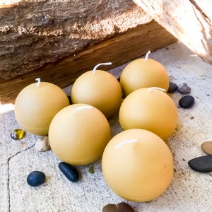 May include: Six round, golden yellow candles with white wicks. The candles are arranged in a circle on a gray surface.