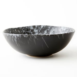 Puede incluir: Un cuenco de cerámica negro con un esmalte blanco moteado. El cuenco tiene un borde ancho y un fondo redondeado.