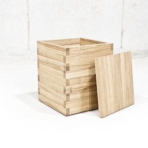 Puede incluir: Una caja de almacenamiento de madera con tapa. La caja está hecha de madera clara con un patrón de veta natural. La tapa está separada de la caja y también está hecha de madera clara.