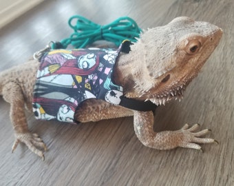 May include: A bearded dragon wearing a black and white harness with a Nightmare Before Christmas print. The harness has a teal leash attached to it.