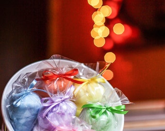 May include: A white bowl filled with six colorful, wrapped bath bombs. Each bath bomb is a different pastel color: blue, pink, purple, green, yellow, and orange. Each is tied with a matching ribbon. The background is a blurred red with bokeh lights.