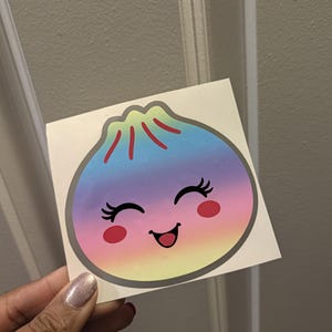 Könnte beinhalten: Ein quadratischer Aufkleber mit einem Cartoon-Knödel, der einen Farbverlauf von Blau oben zu Pink und Gelb unten aufweist. Der Knödel hat ein lächelndes Gesicht mit geschlossenen Augen, langen Wimpern und roten Wangen.