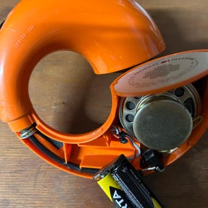 May include: A bright orange, circular National Panasonic radio with its casing partially open, revealing an internal speaker, wiring, and a single Varta Super Heavy Duty AA battery. The device features a curved design with the text OFF-VOLUME visible.
