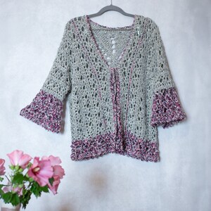 Könnte beinhalten: Ein gehäkeltes Cardigan in Grau und Rosa. Das Cardigan hat einen V-Ausschnitt und lange Ärmel mit einer gehäkelten Bordüre.