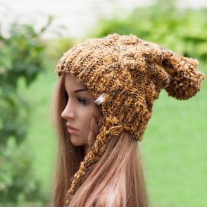 Può includere: Un berretto di lana marrone e nero con una lunga coda pendente. Il berretto ha un pon pon in cima e un piccolo cartellino bianco con la scritta "Felt" su di esso.