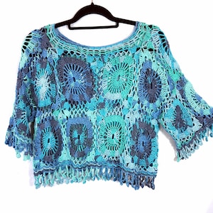 May include: A crochet top featuring a pattern of circular motifs in shades of dark blue, medium blue, and turquoise. The garment has a wide neckline, three-quarter length sleeves, and a bottom hem finished with a fringe of hanging yarn loops.