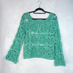 Könnte beinhalten: Ein grüner Häkelpullover mit lockerer Passform und Glockenärmeln. Der Pullover ist aus einem leichten, offen gewebten Stoff.
