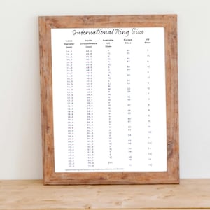 May include: A wooden desk with a framed chart labeled "International Ring Size." A brown water bottle, a candle in a glass, and two notebooks are on the desk. A wooden chair and a blank white paper are in the foreground.