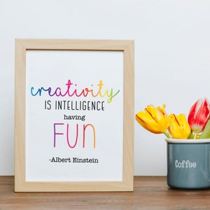 May include: A framed print with a quote by Albert Einstein: "Creativity is intelligence having fun." The quote is written in rainbow colours and the frame is light wood.