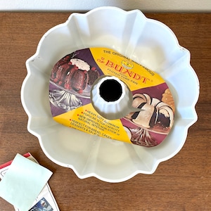 Könnte beinhalten: Eine weiße, geriffelte Bundt-Kuchenform mit einem Fassungsvermögen von 12 Tassen. Die Form hat ein zentrales Rohr und einen gewellten Rand. Die Originalverpackung befindet sich in der Form mit dem Text "The Original and Famous Bundt".