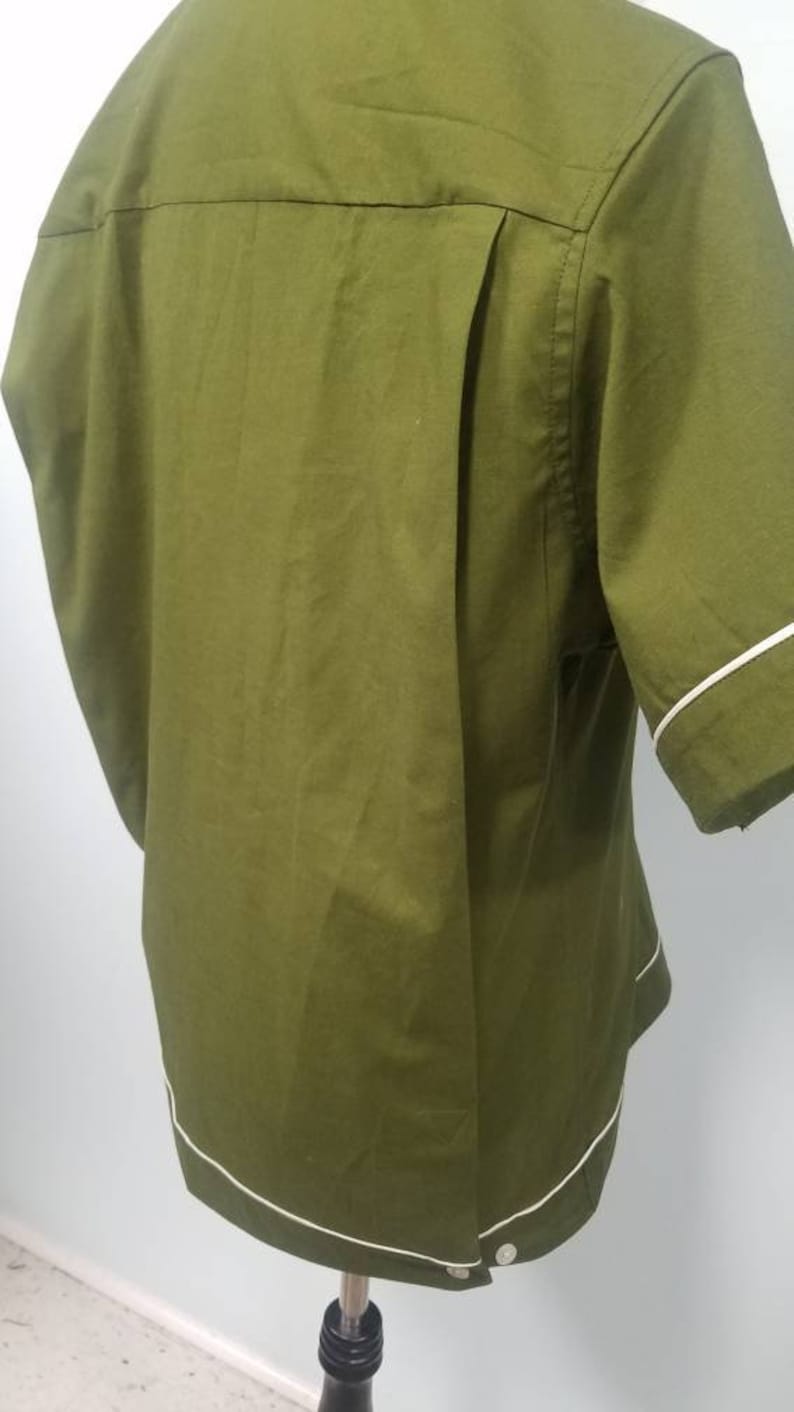 May include: Olive green short-sleeved shirt with white trim on the sleeves and hem. The back of the shirt features a pleated design. The shirt is buttoned at the bottom and is displayed on a mannequin.