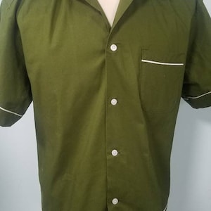 May include: Olive green short-sleeved shirt with a camp collar and white trim. The shirt has a button-down front, a chest pocket, and white trim along the cuffs and hem. The shirt is displayed on a mannequin.