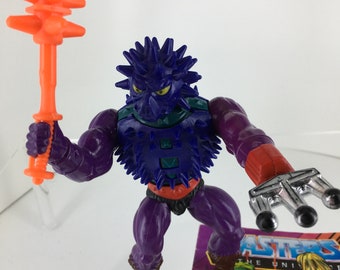 May include: Purple action figure with a spiky head and a spiked chest piece. The figure is holding an orange spiked weapon in one hand and a silver claw in the other. The figure is standing on a comic book with the text "Masters of the Universe" on it.