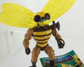 May include: A vintage Masters of the Universe action figure of Buzz-Off, a bee-like character with yellow wings, brown and yellow armor, and a black axe. The figure is standing on a comic book featuring He-Man and other Masters of the Universe characters.