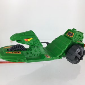 May include: Green plastic toy vehicle with a large, detailed, green, snake-like head with an orange eye. The vehicle has a large, black wheel and a smaller, orange wheel. The vehicle has a green, plastic body with a black, plastic wheel.